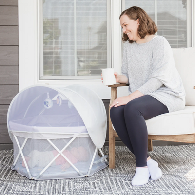 Mom outside on deck watching baby lay in My Play Baby Portable Infant Bassinet.