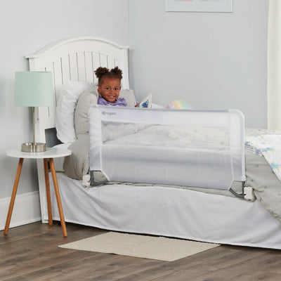 A child sitting in a bed with a white swing down bed rail installed on the side. The bed is placed in a room with a nightstand and a lamp.
