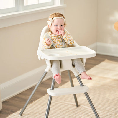 A baby sitting in a white Regalo Baby Basics High Chair with a plastic and steel construction, accompanied by an attachable tray, in a home setting.