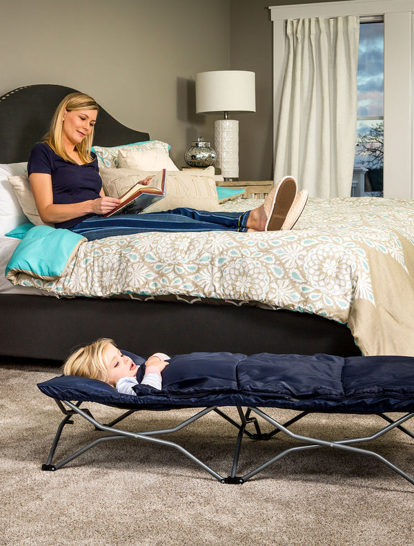 Child sleeps on the Deluxe My Cot® Portable Toddler Travel Bed™ while parent reads